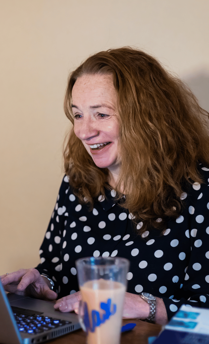 Maxine smiles at a laptop. A cup of coffee is in shot in a glass mug with 'Max' written on. She is wearing a black top with white polka dots.