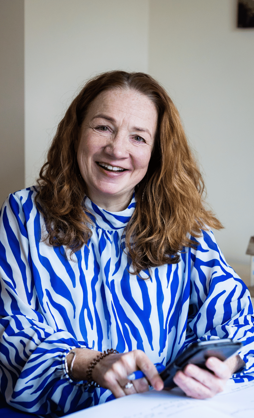 Maxine smiles at the camera, with curly red hair and a blue and white shirt. She is holding a mobile phone.