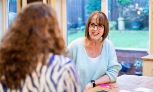 A woman with short brown hair and glasses leans on a desk and smiles at Maxine - you can only see the back of Maxines head with her red hair.